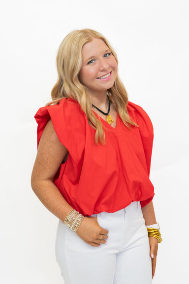 A smiling blonde woman wearing a bright red sleeveless top with voluminous gathered shoulders and white pants, standing against a plain white studio background.