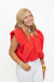 A smiling blonde woman wearing a bright red sleeveless top with voluminous gathered shoulders and white pants, standing against a plain white studio background.