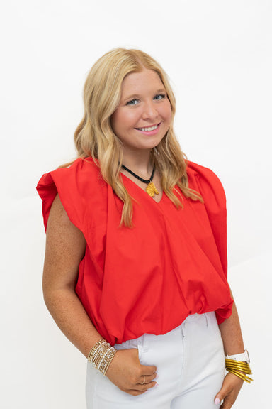 A smiling blonde woman wearing a bright red sleeveless top with puffed shoulders and white pants, accessorized with gold bracelets and a necklace, photographed against a plain white studio background.