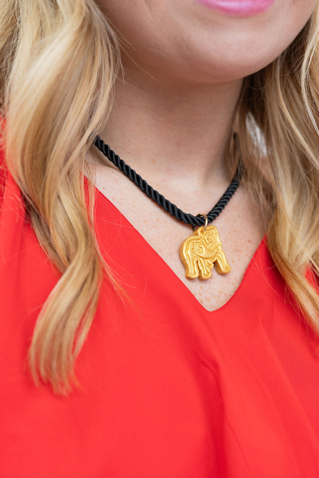 A close-up of a woman wearing a black braided cord necklace with a small gold elephant pendant, paired with a bright red top.