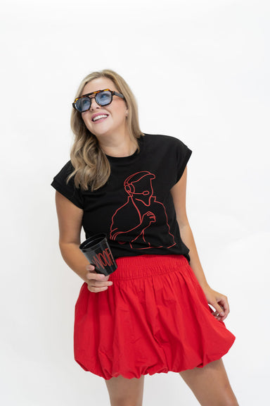 A woman modeling a black graphic t-shirt with a red line art design and a bright red pleated mini skirt, wearing tortoiseshell sunglasses and holding a black coffee cup with red text, set against a plain white studio background.