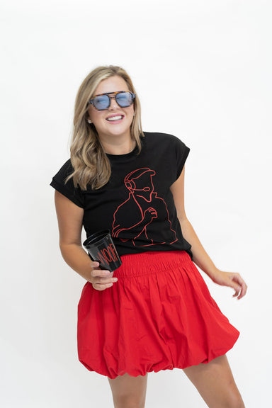A woman modeling a black graphic t-shirt with a red line art design and a bright red pleated mini skirt, wearing blue-tinted sunglasses and holding a black coffee cup against a plain white background.