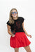 A woman modeling a black graphic t-shirt with a red line art design and a bright red pleated mini skirt, wearing blue-tinted sunglasses and holding a black coffee cup against a plain white background.