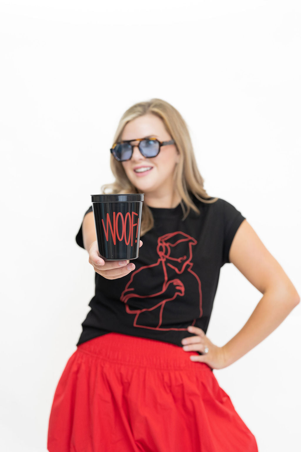 A woman wearing black sunglasses, a black graphic t-shirt with red line art, and a red skirt holds up a black cup with the word 'WOOF' printed in red, set against a plain white background.