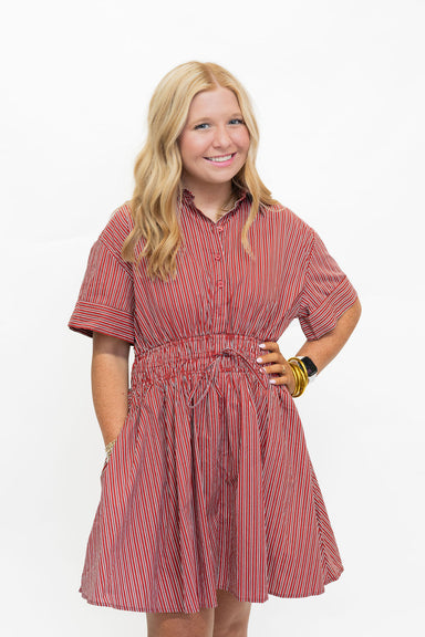A young blonde girl modeling a short-sleeve red and white vertically striped button-down dress with a smocked waist and side pockets, posed against a solid white background.