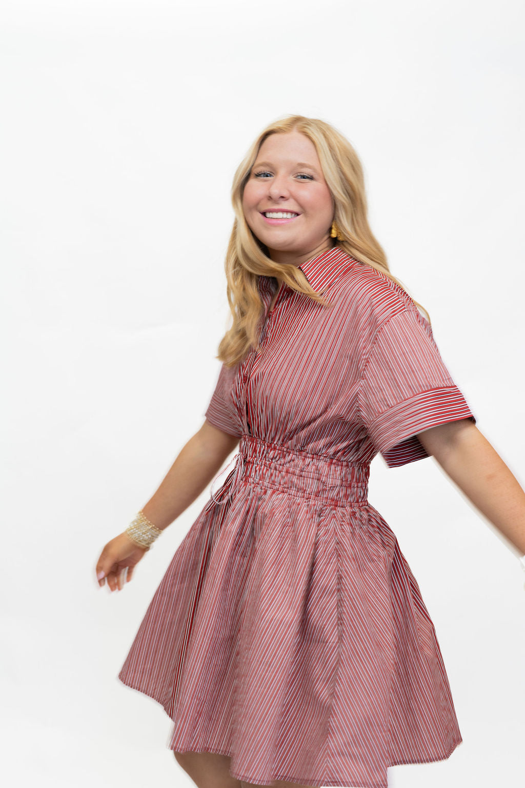 A smiling blonde woman wearing a short-sleeve pink and red vertically striped dress with a smocked waist, posed against a solid white background.