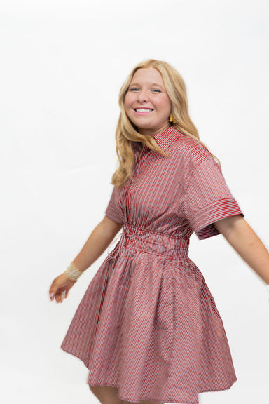 A smiling blonde woman wearing a short-sleeve pink and red vertically striped dress with a smocked waist, posed against a solid white background.