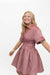A smiling blonde woman wearing a short-sleeve pink and red vertically striped dress with a smocked waist, posed against a solid white background.