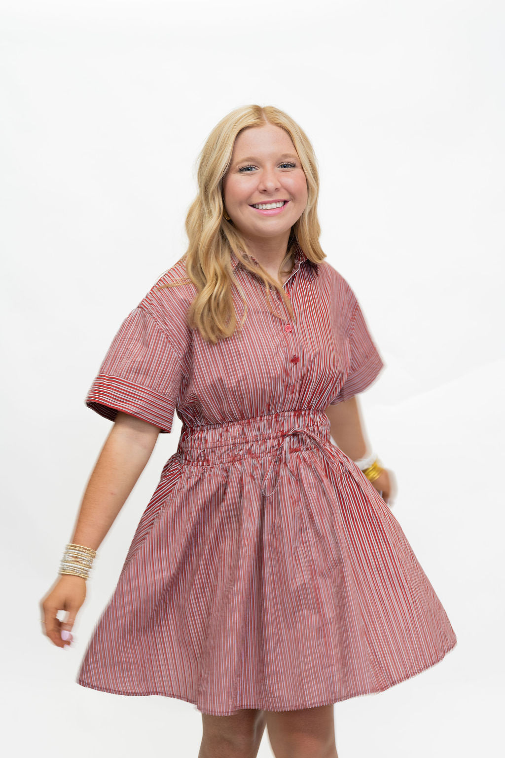 A smiling blonde woman wearing a short-sleeve pink and white vertically striped button-up dress with a gathered waist, standing against a plain white background.