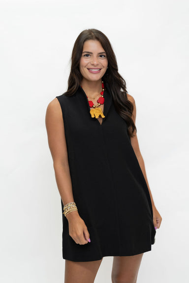 A woman modeling a sleeveless black V-neck mini dress with a small front slit, paired with a red and yellow statement necklace and gold bracelets, against a plain white studio background.