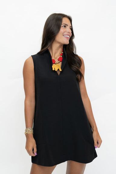A woman modeling a sleeveless black shift dress with a notched lapel collar, paired with a red and yellow statement necklace, against a plain white studio background.