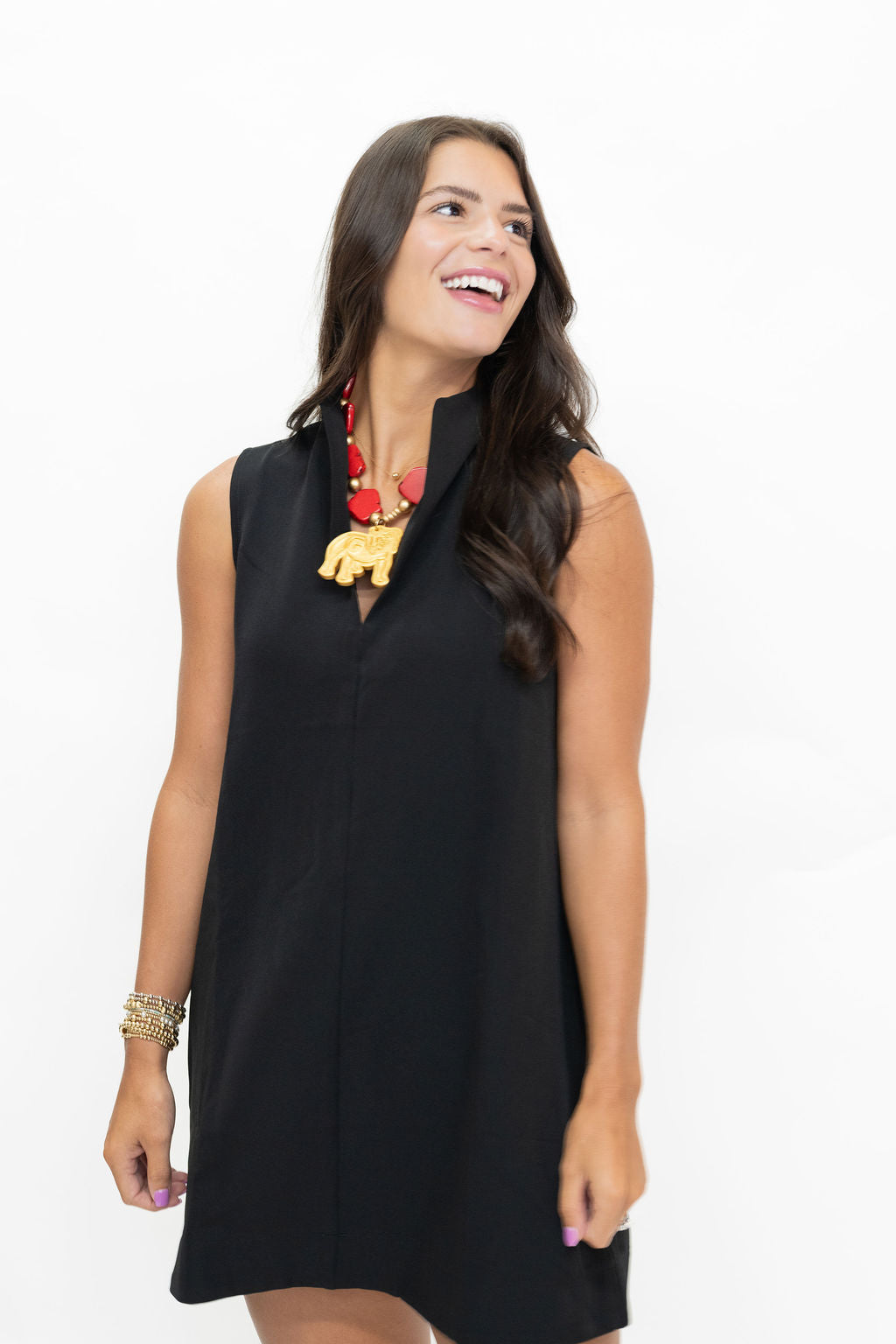 A woman wearing a sleeveless black shift dress with a split neckline and a gold elephant pendant necklace, posing against a solid white background.