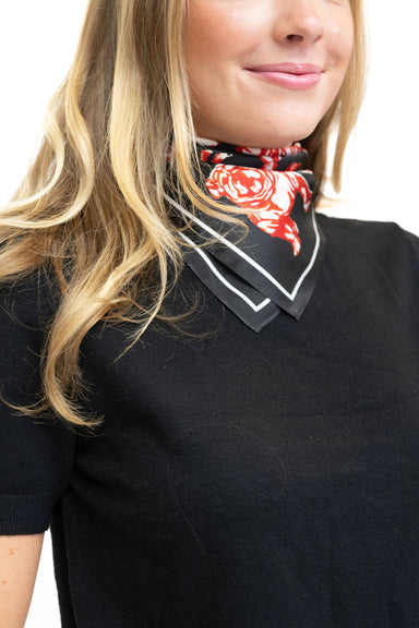 A close-up of a woman wearing a black short-sleeve top and a black neck scarf with a red rose pattern and white border trim, set against a plain white background.