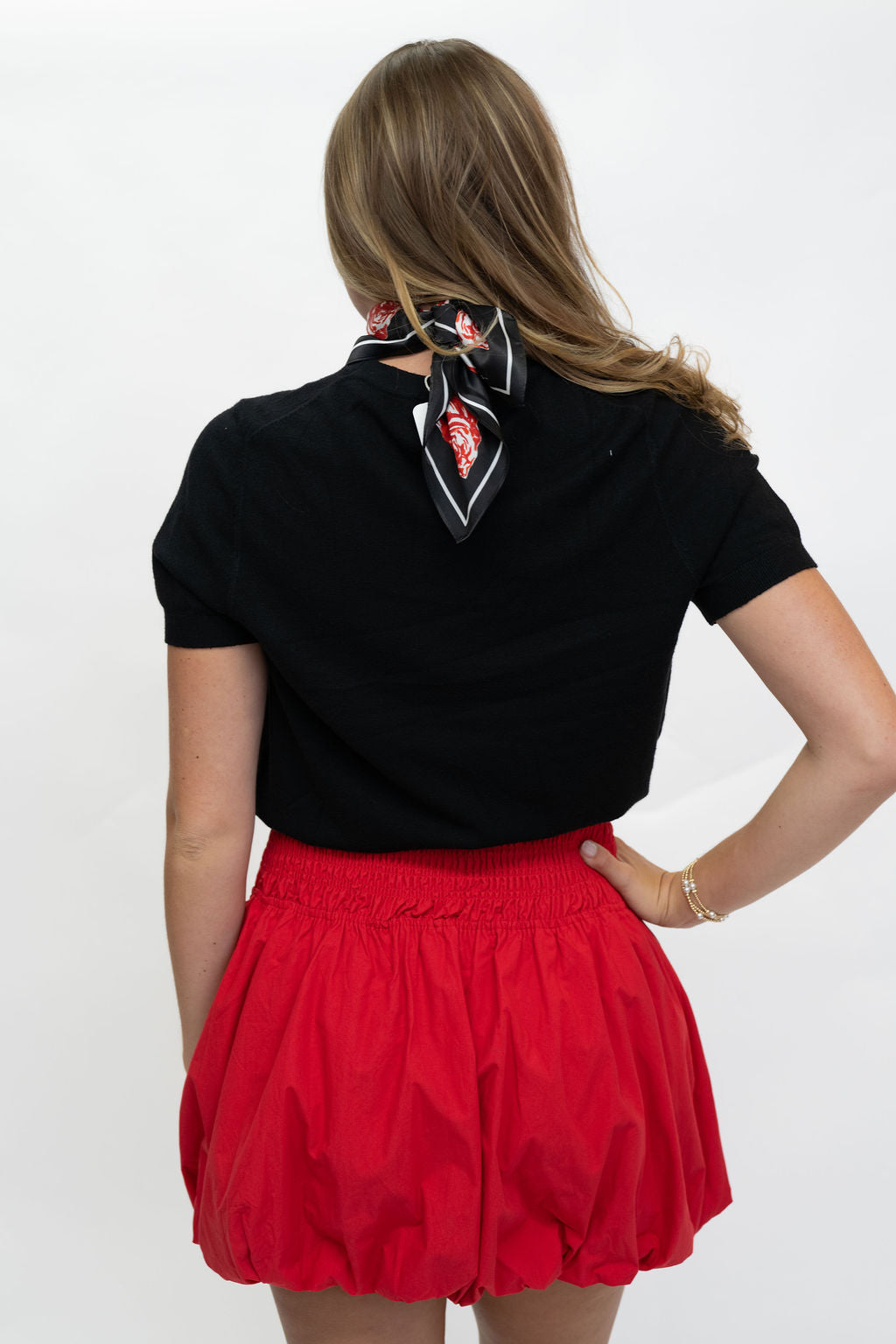 A woman shown from the back wearing a black short-sleeve top with a red, black, and white patterned neck scarf, paired with a red pleated skirt, against a plain white background.