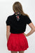 A woman shown from the back wearing a black short-sleeve top with a red, black, and white patterned neck scarf, paired with a red pleated skirt, against a plain white background.