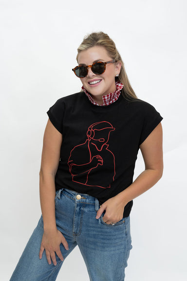 A woman wearing a black short-sleeve t-shirt with a red line art graphic of a person, styled with blue jeans, a red and white checkered neck gaiter, and sunglasses, against a plain white studio background.