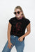 A woman wearing a black short-sleeve t-shirt with a red line art graphic of a person, styled with blue jeans, a red and white checkered neck gaiter, and sunglasses, against a plain white studio background.