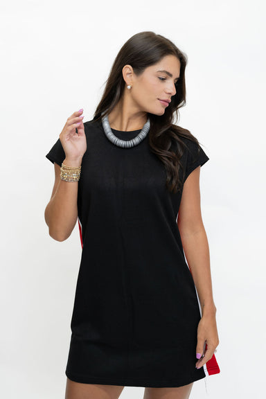 A woman modeling a black short-sleeve shift dress with a textured gray statement necklace and gold bracelets, posed against a plain white studio background.