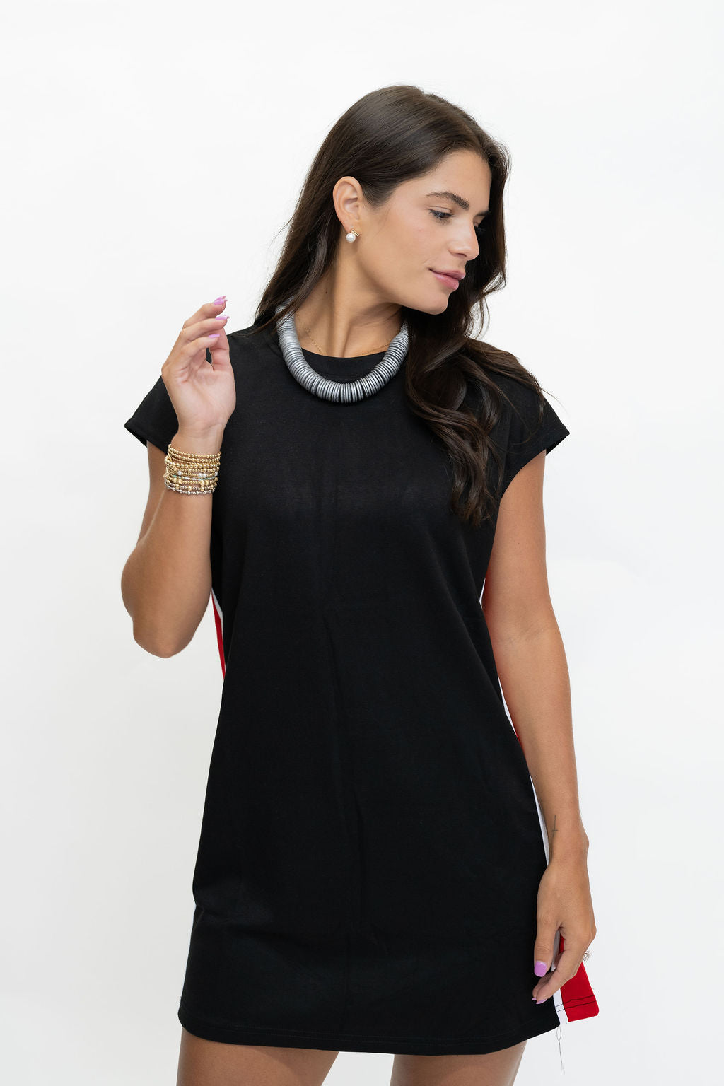 A woman modeling a black short-sleeve shift dress with a textured gray statement necklace and gold bracelets, posed against a plain white studio background.