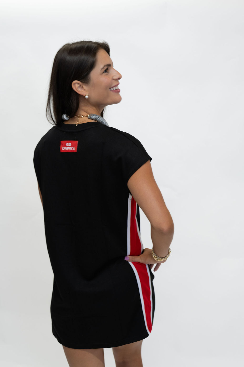 A woman wearing a black short-sleeve mini dress with red and white side stripes and a red square logo patch on the back, photographed against a plain white studio background.