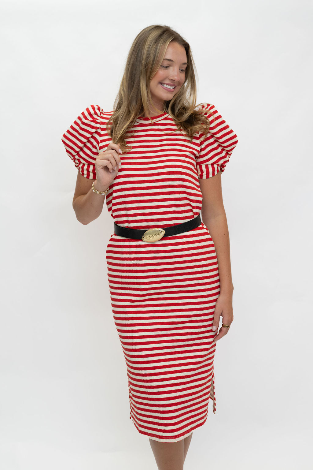 A woman modeling a red and white horizontally striped dress with puffed short sleeves, a black belt with a gold oval buckle, and a side slit with tassels, photographed against a plain white studio background.