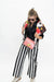 A woman modeling a black and white vertically striped wide-leg pant set, a black top, a colorful patterned cardigan, and a red-and-white gingham crossbody bag with a gold clasp, against a plain white studio background.