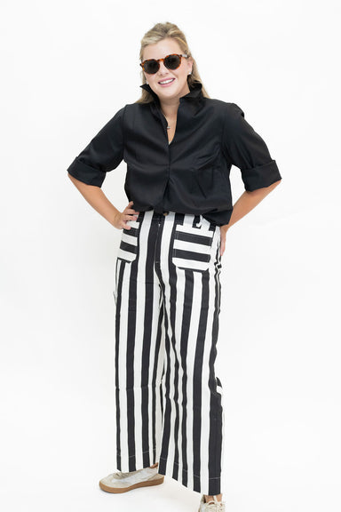 A woman modeling a black button-up shirt and black-and-white vertically striped wide-leg pants, styled with sunglasses and beige sneakers against a plain white studio background.
