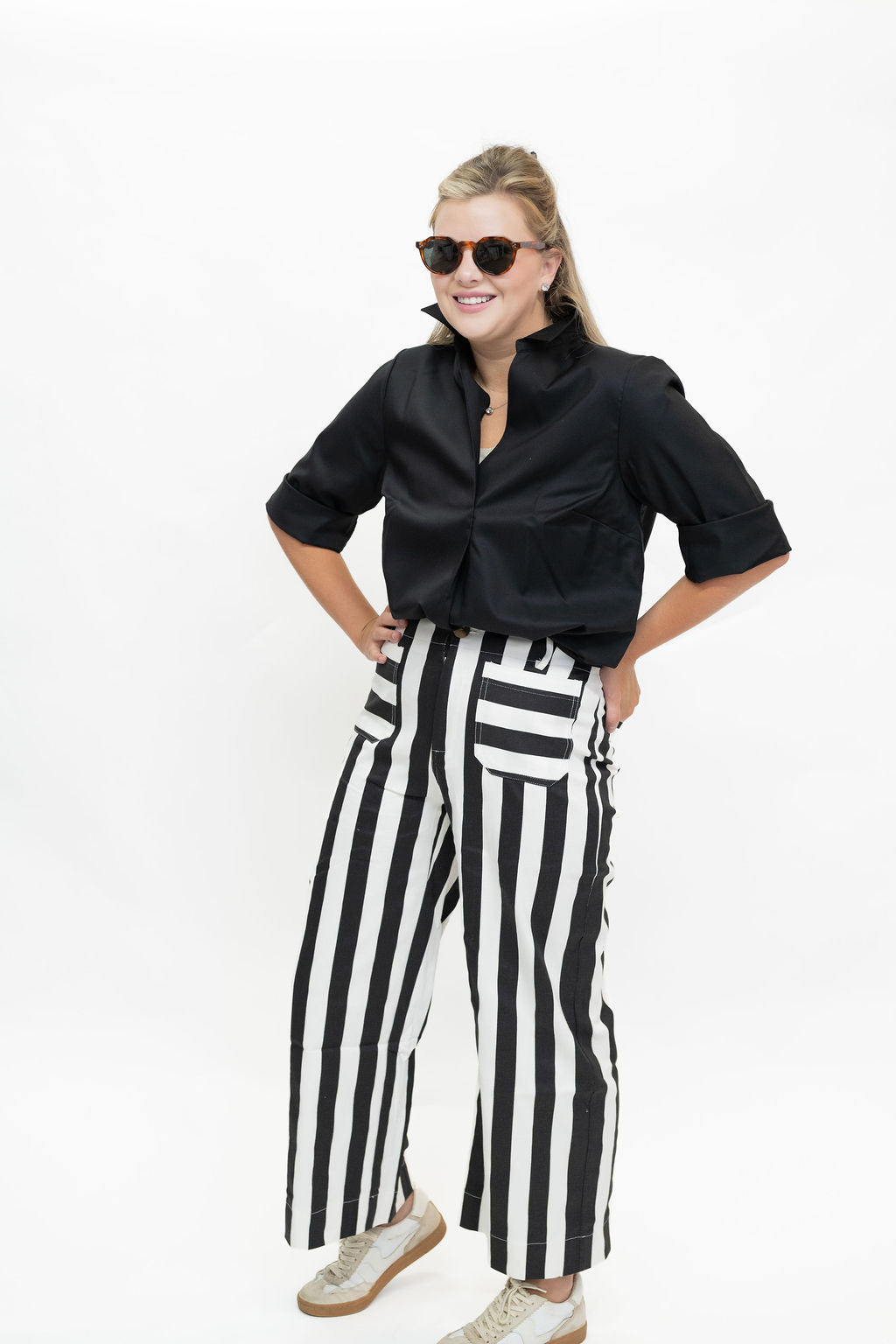 A woman modeling a black satin short-sleeve button-up shirt and high-waisted black and white vertically striped wide-leg pants, styled with beige sneakers and sunglasses, against a plain white background.