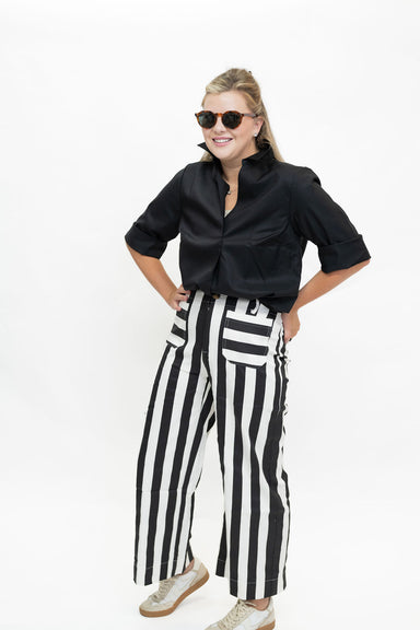 A woman modeling a black satin short-sleeve button-up shirt and high-waisted black and white vertically striped wide-leg pants, styled with beige sneakers and sunglasses, against a plain white background.