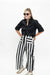 A woman modeling a black satin short-sleeve button-up shirt and high-waisted black and white vertically striped wide-leg pants, styled with beige sneakers and sunglasses, against a plain white background.