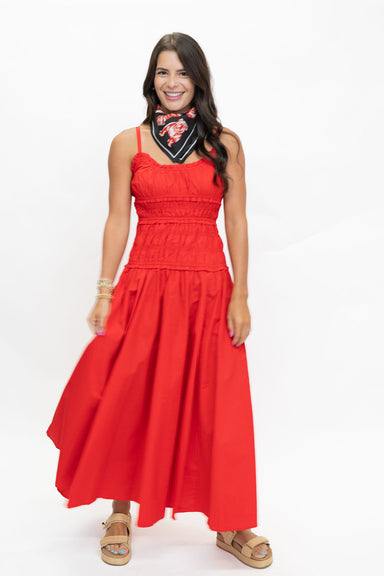 A woman modeling a bright red sleeveless maxi dress with a smocked bodice and flowing skirt, paired with a black bandana and tan sandals, against a plain white studio background.