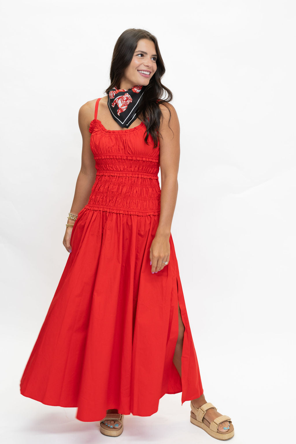 A woman modeling a bright red sleeveless maxi dress with a smocked bodice, thin straps, and a high side slit. She is wearing a black bandana with red and white floral details and tan sandals, standing against a plain white studio background.