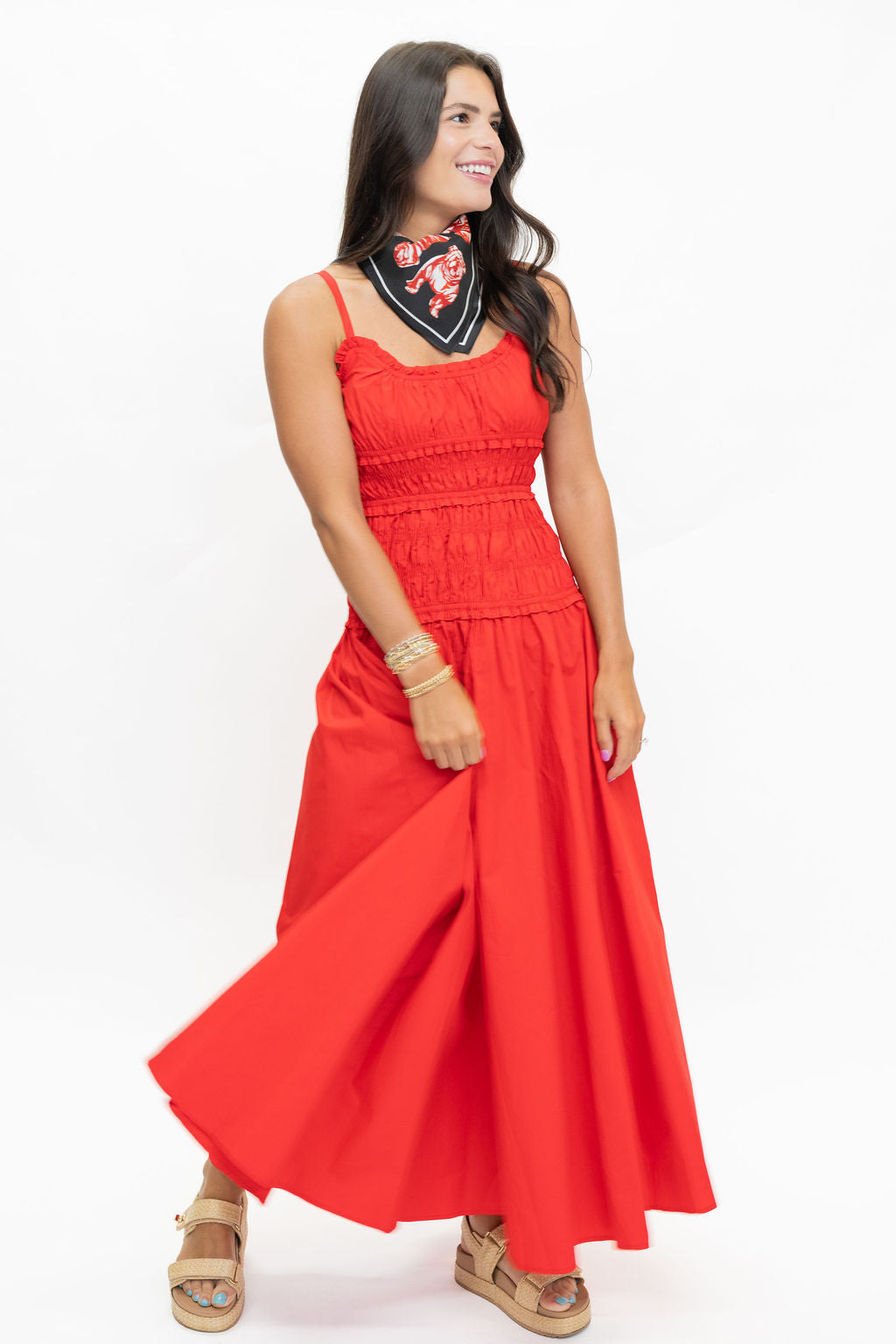 A woman modeling a bright red sleeveless maxi dress with a smocked bodice and a matching red skirt, paired with a black bandana featuring a red floral pattern, on a white studio background.
