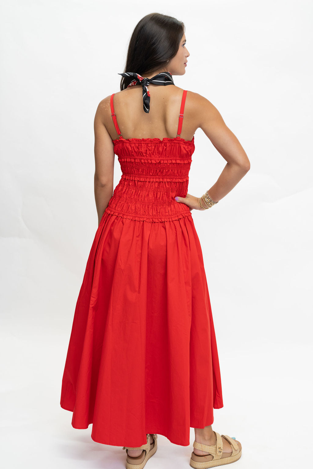 A woman wearing a bright red sleeveless maxi dress with thin straps, a smocked bodice, and a gathered skirt, shown from the back against a plain white studio background.