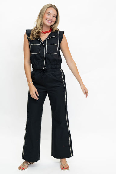 A woman modeling a black sleeveless jumpsuit with white contrast stitching, wide-leg pants, and a drawstring waist, set against a plain white studio background.