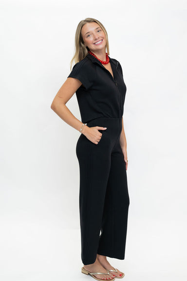A woman modeling a black short-sleeve collared jumpsuit with wide legs, paired with a red beaded necklace and gold sandals, against a plain white studio background.