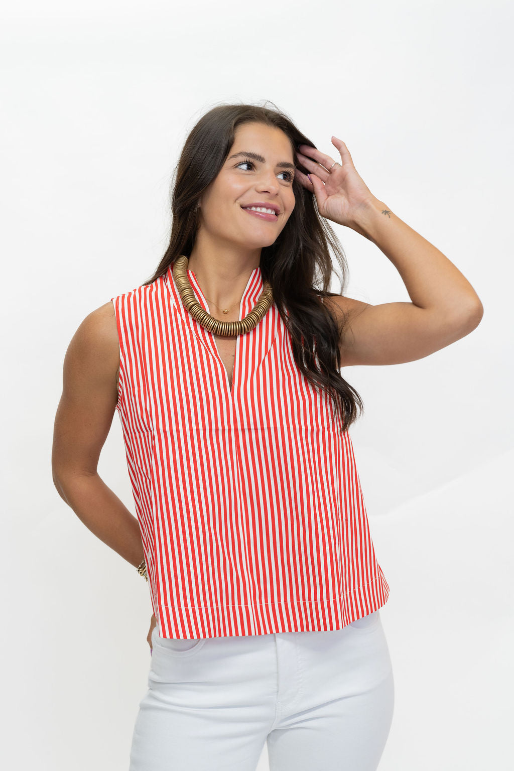 A woman wearing a sleeveless red and white vertically striped top with a high collar, paired with white pants and a chunky gold necklace, posing against a plain white background.