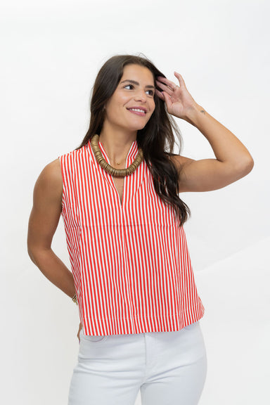 A woman wearing a sleeveless red and white vertically striped top with a high collar, paired with white pants and a chunky gold necklace, posing against a plain white background.