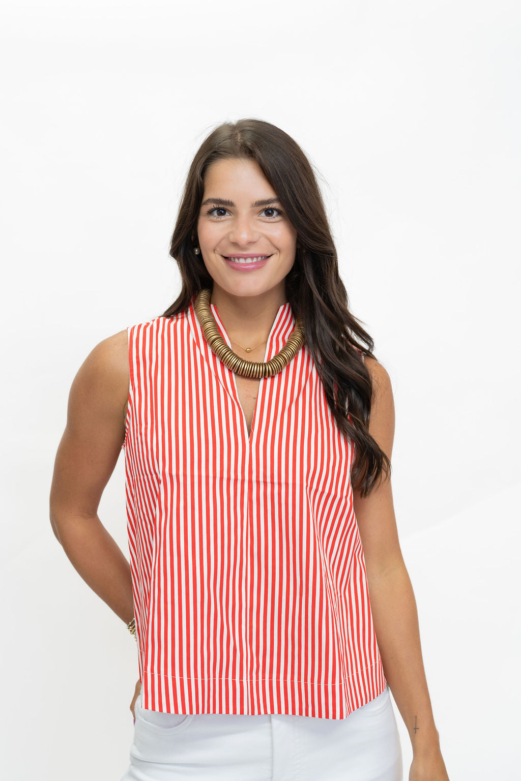 A woman modeling a sleeveless top with vertical red and white stripes, paired with white pants and a chunky gold necklace, against a plain white background.