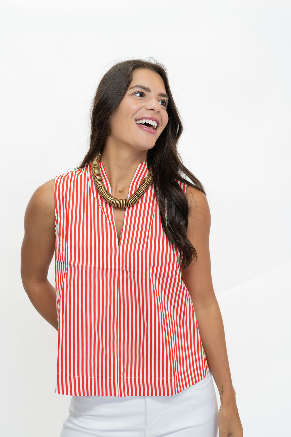 A woman wearing a sleeveless red and white vertically striped top with a split V-neckline, paired with white pants and a chunky gold necklace, posing against a solid white background.