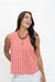 A woman wearing a sleeveless red and white vertically striped top with a split V-neckline, paired with white pants and a chunky gold necklace, posing against a solid white background.