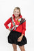 A woman posing in a bright red long-sleeve collared shirt with a black, white, and red patterned scarf tied around the collar, paired with a black pleated mini skirt, against a plain white background.