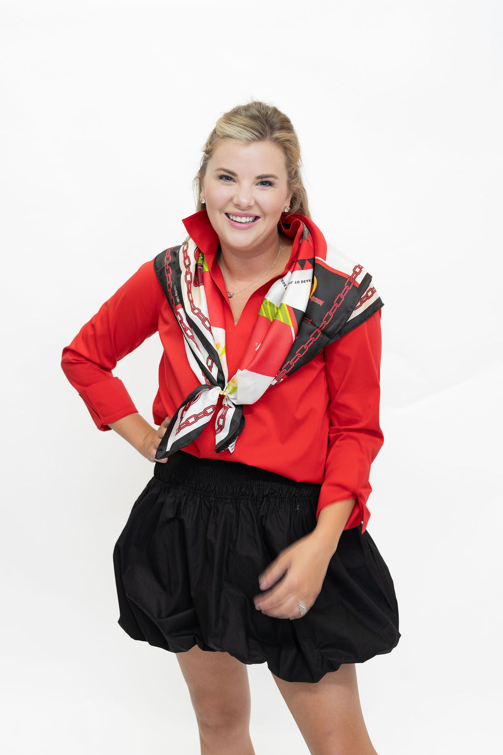 A woman posing in a bright red long-sleeve collared shirt with a black, white, and red patterned scarf tied around the collar, paired with a black pleated mini skirt, against a plain white background.