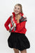 A woman posing in a red long-sleeve collared shirt, a black pleated mini skirt, and a black, white, and red patterned scarf, set against a plain white studio background.