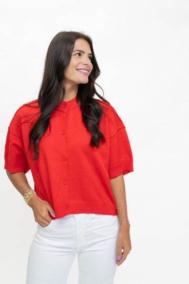 A woman wearing a bright red short-sleeve button-up blouse with structured shoulders and white pants, standing against a plain white studio background.