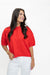 A woman wearing a bright red short-sleeve button-up blouse with structured shoulders and white pants, standing against a plain white studio background.