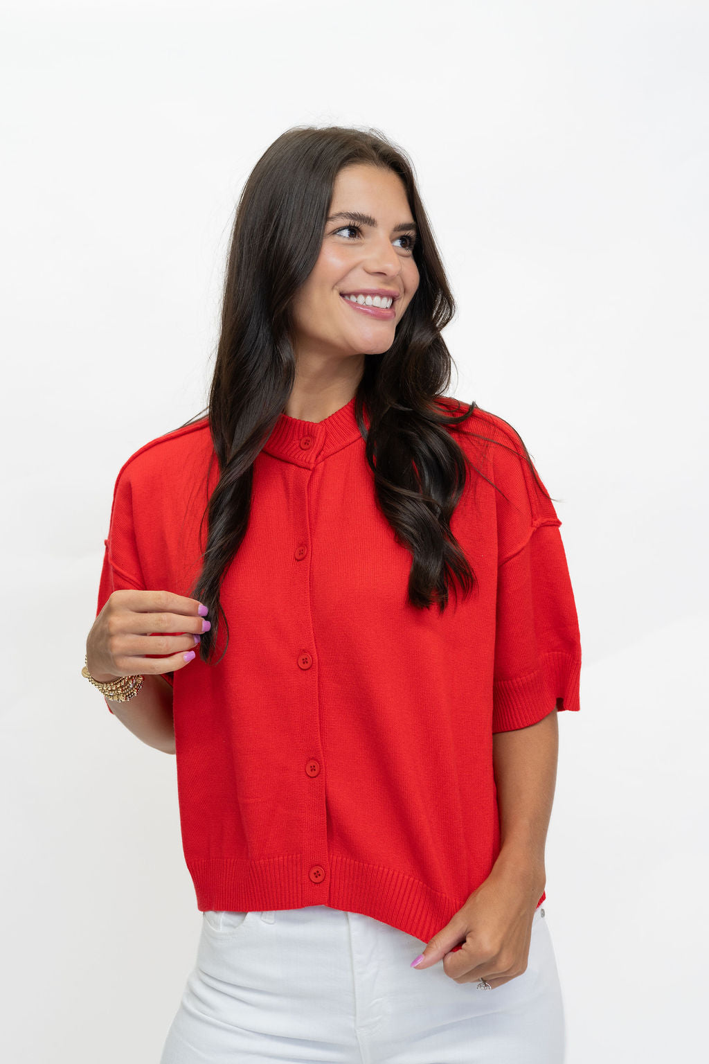 A woman with long dark hair wearing a bright red short-sleeve knit cardigan top and white pants, standing against a plain white studio background.