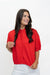 A woman with long dark hair wearing a bright red short-sleeve knit cardigan top and white pants, standing against a plain white studio background.