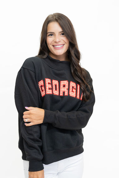 A woman wearing a black crewneck sweatshirt with 'GEORGIA' printed across the chest in red block letters outlined in white, paired with white pants, against a plain white background.
