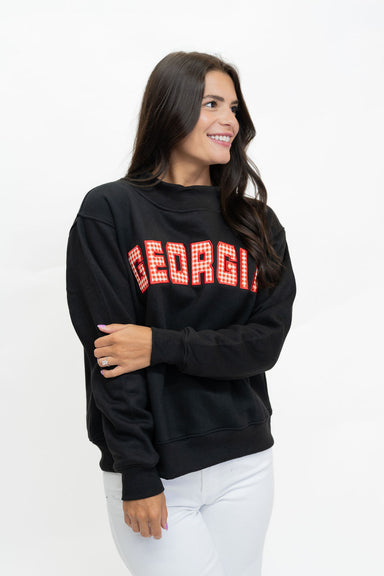 A woman wearing a black crewneck sweatshirt with 'GEORGIA' in red and white gingham block letters across the chest, paired with white pants, photographed against a plain white studio background.